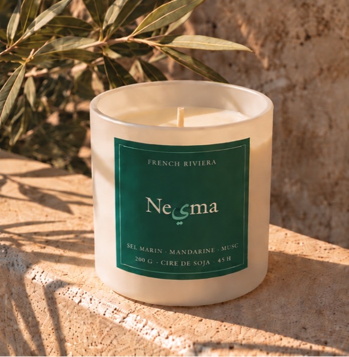 Nesma Aigue Marine candle in matte white glass with its deep green label, on sunlit stone.