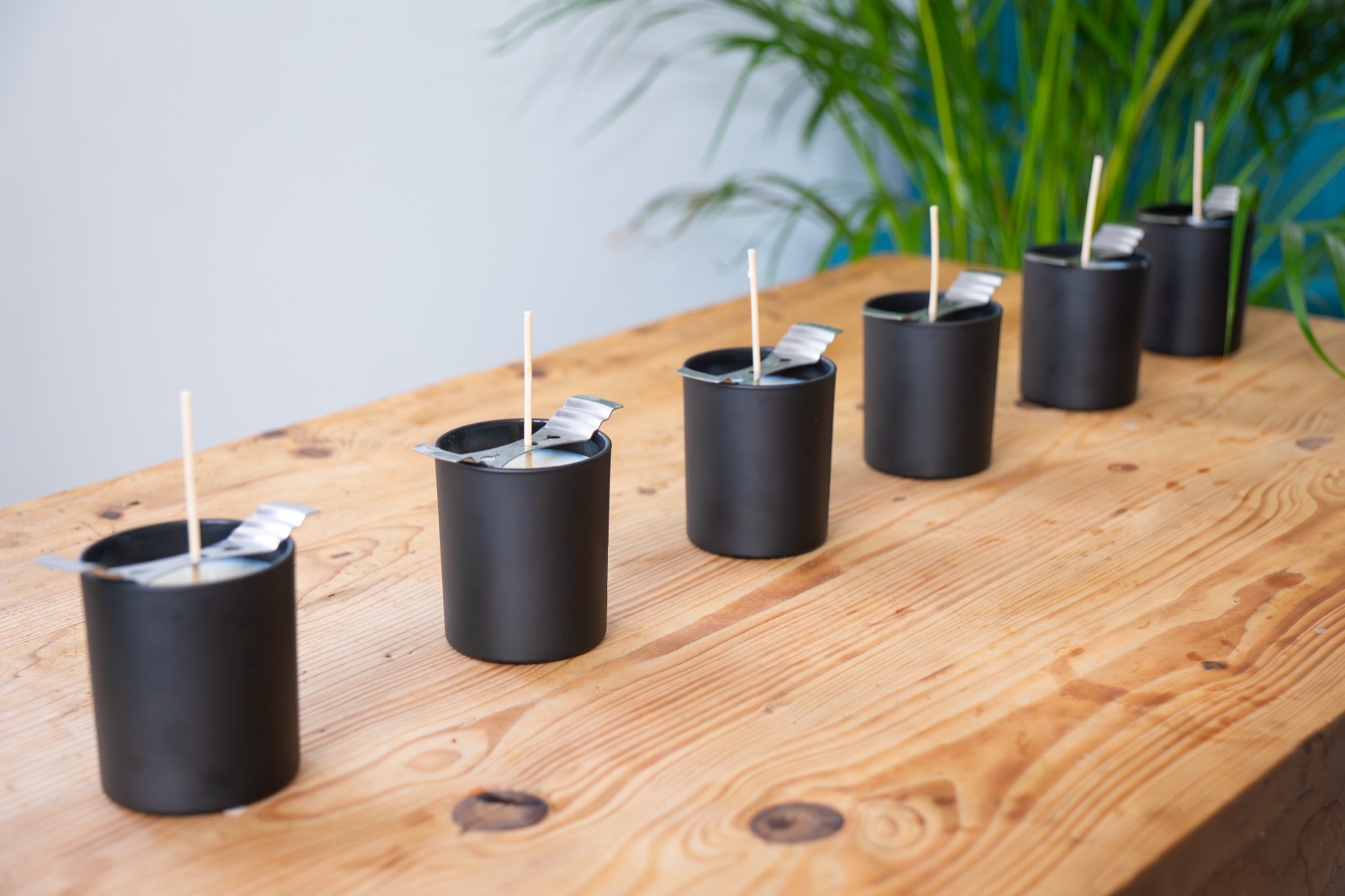 A row of six hand-poured candles in matte black glass, lined up along a wooden table.