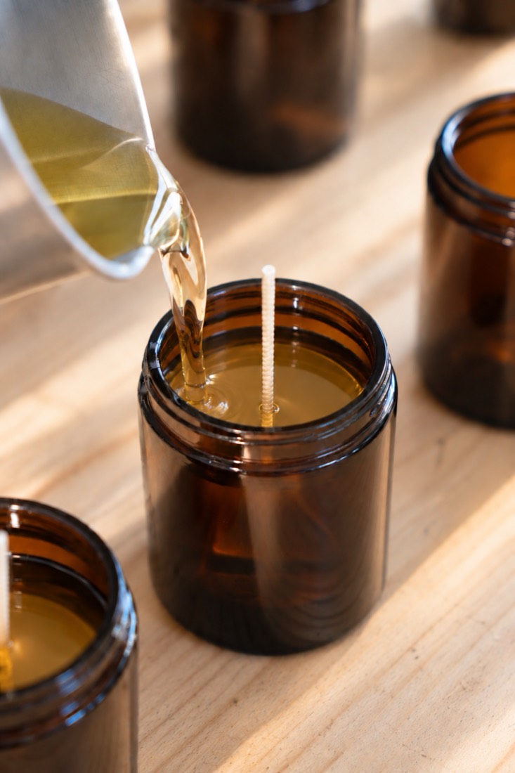 Melted wax being poured from a pitcher into an amber glass jar, around its wick.